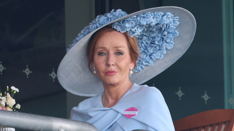 J.K. Rowling wearing a light blue dress with a large ruffled hat with blue flowers and pearl drop earrings