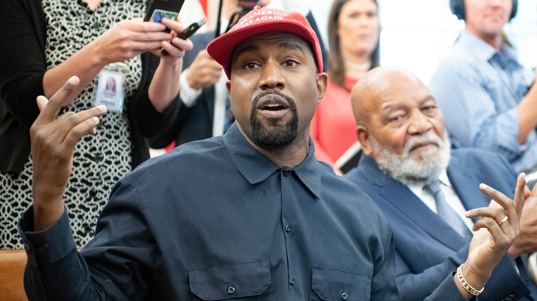 Kanye West speaking with his arms open while wearing a black button down shirt and red Make America Great Again hat