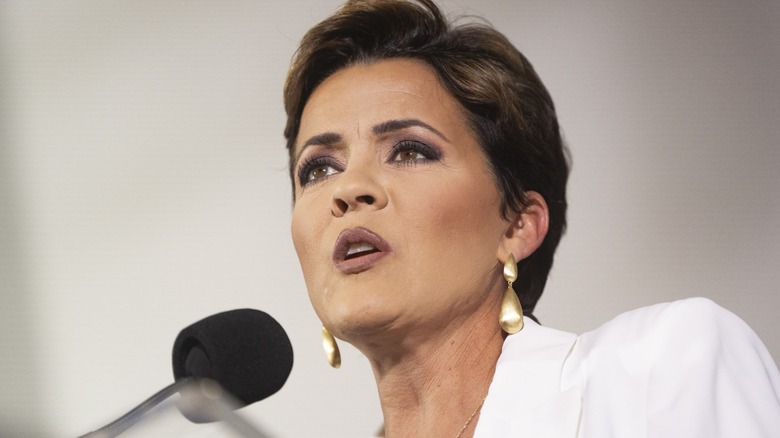 Wearing thick layers of makeup, Kari Laki speaks into a microphone.