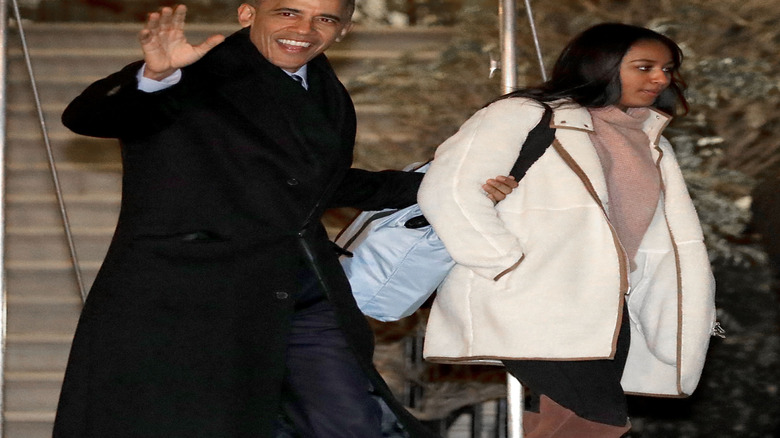 Barack Obama holding the arm of Sasha Obama while waving