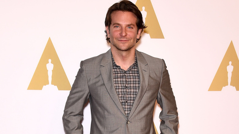 Bradley Cooper smiling with his mouth closed while standing in front of an Oscars background