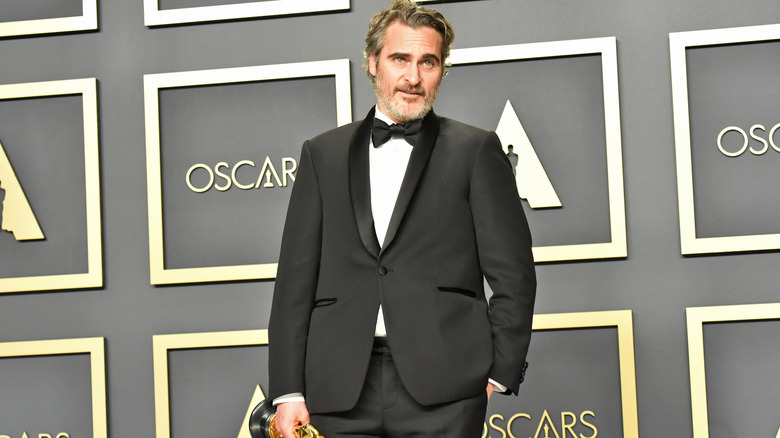 Joaquin Phoenix holding an Oscar statuette looking off camera while posing in front of an Oscars background