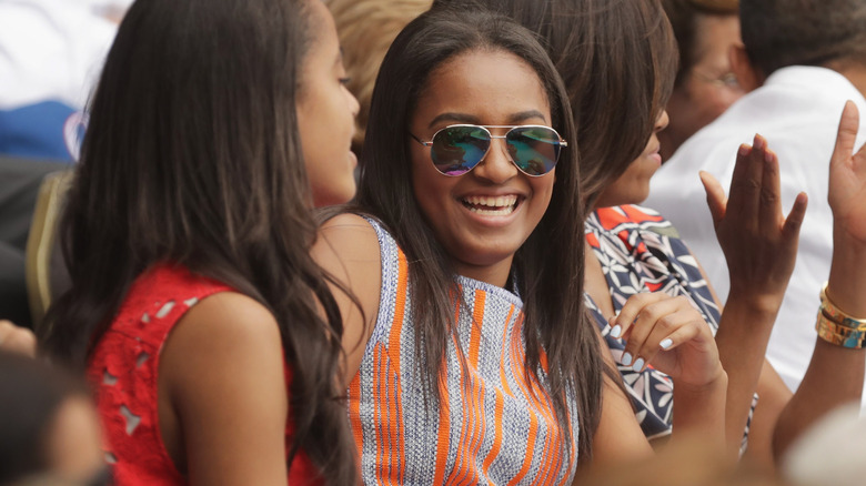 Sasha Obama flashes a wide smile while Michelle Obama claps in the background.