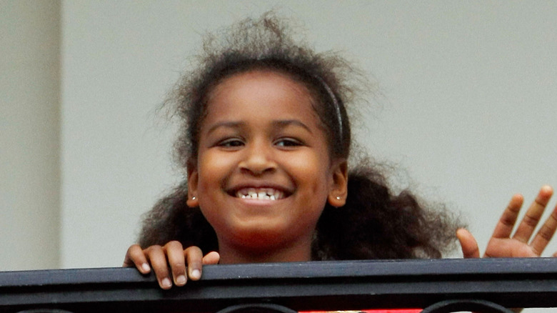 A young Sasha Obama, with many loose strands of hair, flashes a giddy smile from a balcony.