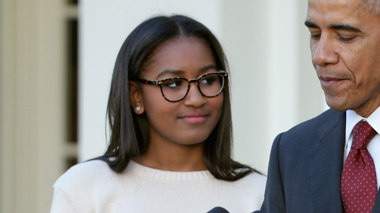Wearing large glasses and minimalist makeup, Sasha Obama looks away from the lens as she stands next to father, Barack Obama..