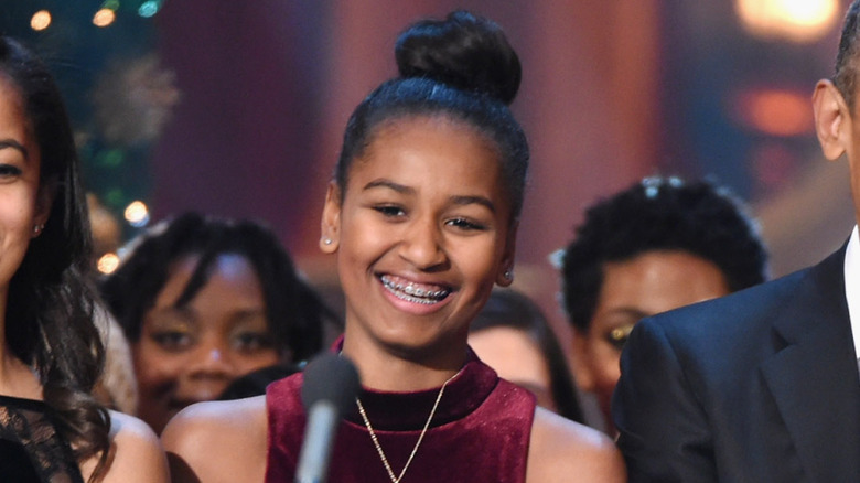 Sasha Obama laughs heartily while standing in front of a microphone.