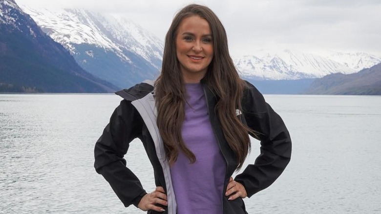 Leah Messer positions her hands on her hips and displays a bright smile as she poses in front of a scenic lake.