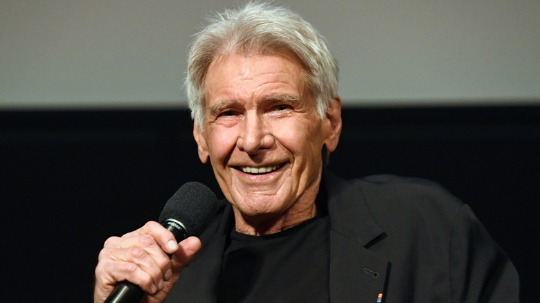 Harrison Ford smiling while holding a mic in an all-black outfit