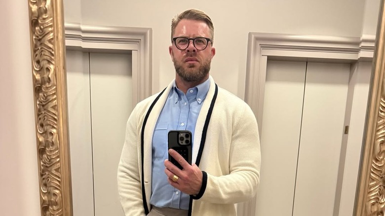 Justin Anderson wearing a blue shirt and cream-colored cardigan and taking a mirror selfie