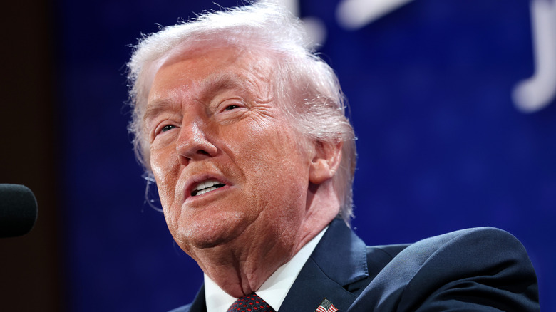 President Donald Trump grimacing while speaking on stage in a navy suit