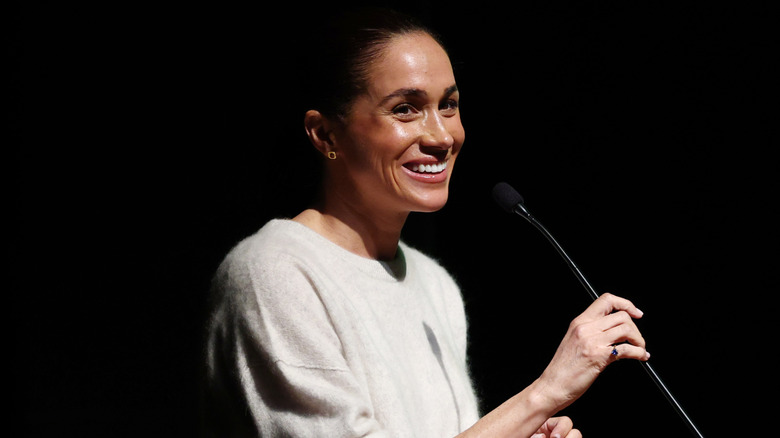 Meghan Markle wears a white sweater while grabbing a microphone.
