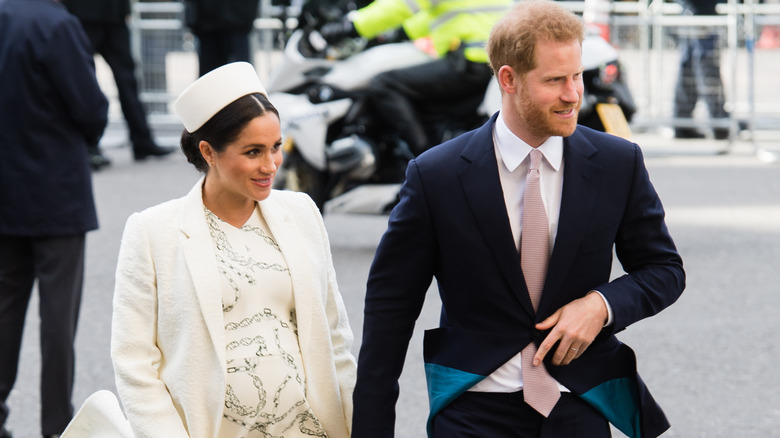 A pregnant Meghan Markle wearing a cream-colored outfit with a matching hat walks beside Prince Harry, who wears a dark blue suit.