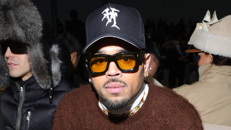 Chris Brown wearing a brown sweater, white turtleneck, black rimmed glasses and a black baseball cap