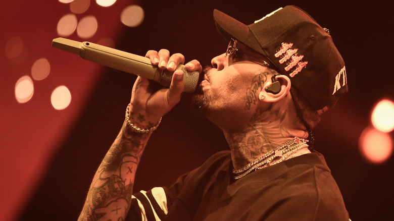 Chris Brown singing into a microphone wearing a black baseball hat, black shirt with white stripes on the sleeve, and silver necklaces and bracelet