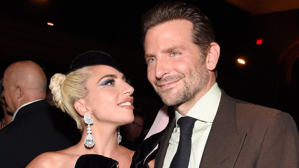 Lady Gaga in a black dress and hat, smiling and looking at a grinning Bradley Cooper