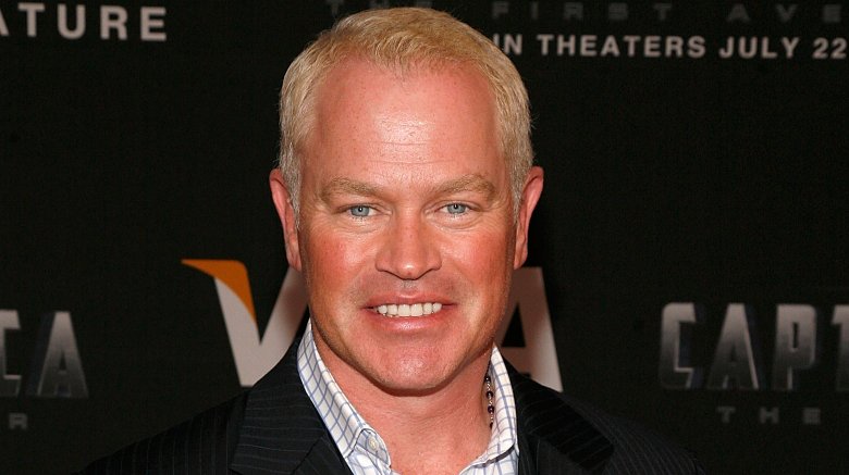Neal McDonough wearing a black pin-striped suit with a white-and-blue checkered shirt, smiling at a movie premiere