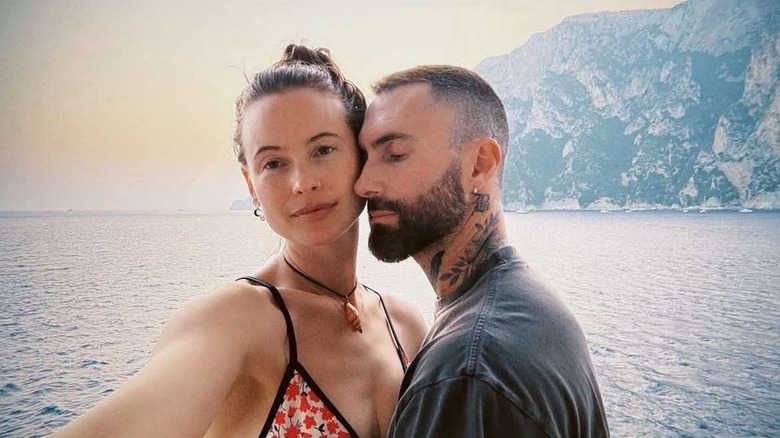 Adam Levine and Behati Prinsloo take a selfie together in a tropical setting with water and mountains in the background.