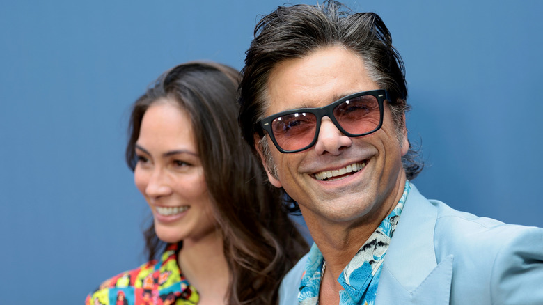 Caitlin McHugh and John Stamos smiling in front of a blue backdrop