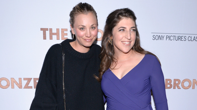 Kaley Cuoco and Mayim Bialik smiling in a black sweater and tight purple dress, respectively.