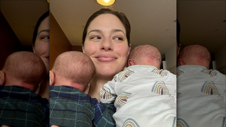 Ashley Graham smiling with two babies on her shoulders