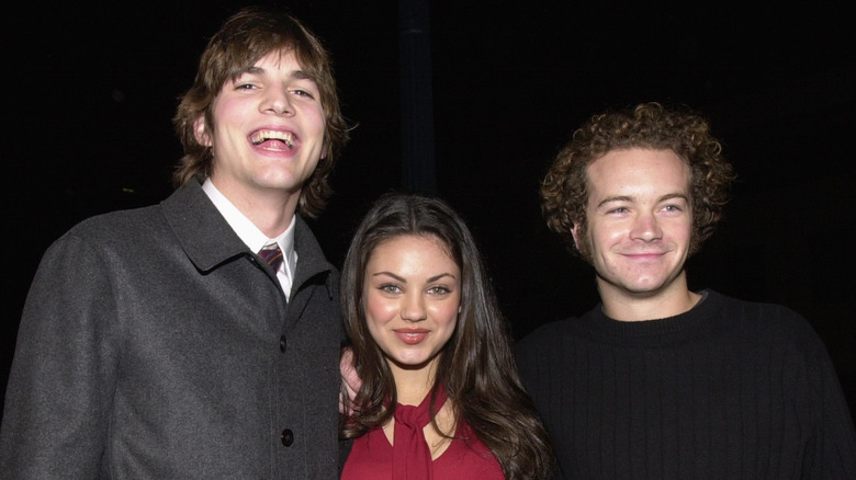 Mila Kunis posing between Ashton Kutcher and Danny Masterson in front of a dark background.