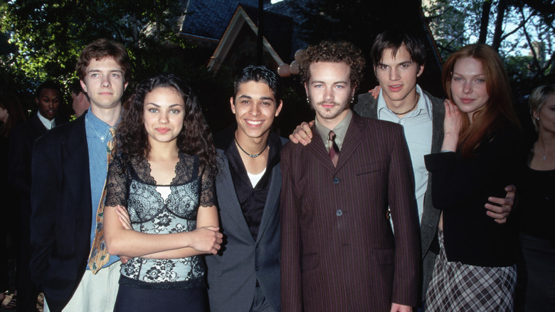 Ashton Kutcher and Mila Kunis with the cast of That '70s Show posing outdoors.