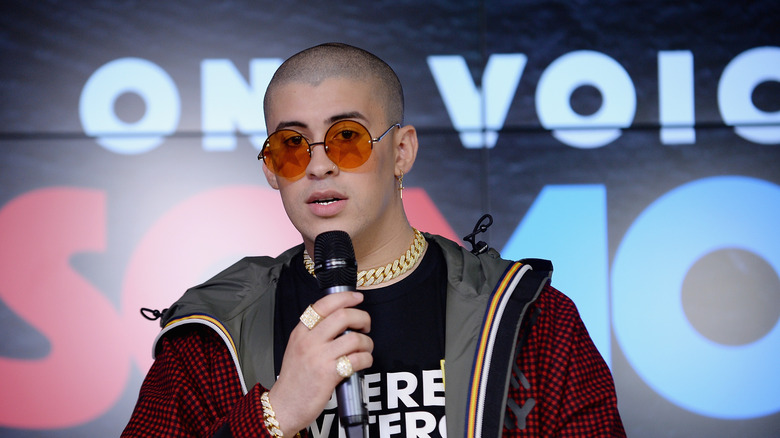 Bad Bunny wearing a red jacket, black t-shirt, and orange-tinted sunglasses while speaking into a microphone