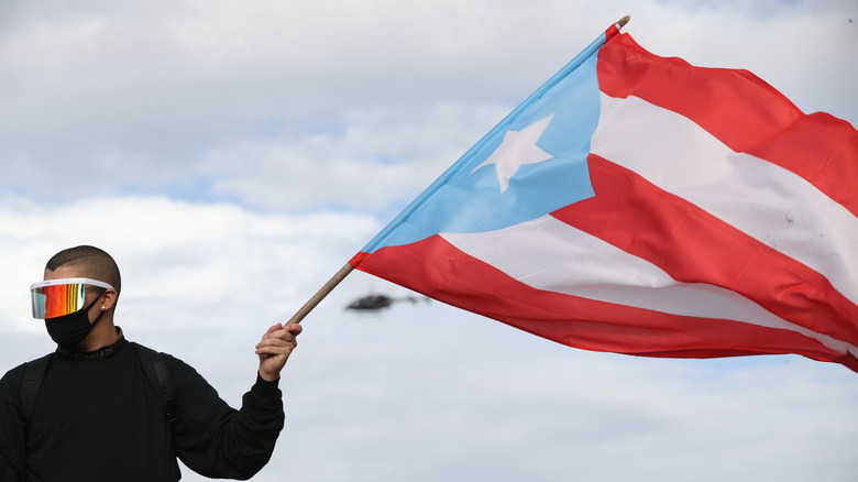 Bad Bunny wearing a black jacket, black mask, and reflective sunglasses while waving the Puerto Rican flag