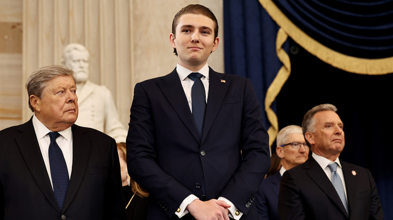 Barron Trump smiling while posing in a dark suit with two much shorter men on either side of him