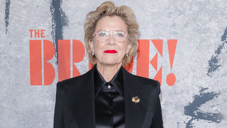 Annette Bening poses in a black jacket and matching shirt with curled blond hair and bright red lipstick.