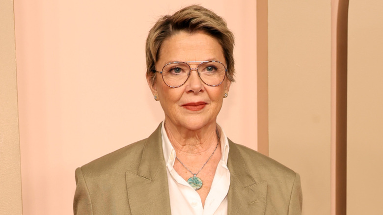 Annette Bening sports a dark blond pixie cut and wears glasses, a green medallion necklace, and a tan blazer.