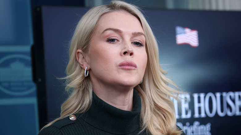 Karoline Leavitt pouting while standing behind the podium in the White House press briefing room in a black turtleneck