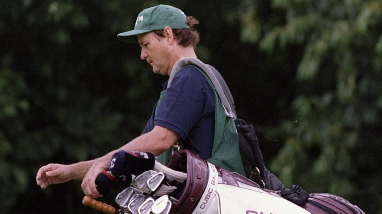 Bill Murray walks along a golf course carrying someone's clubs