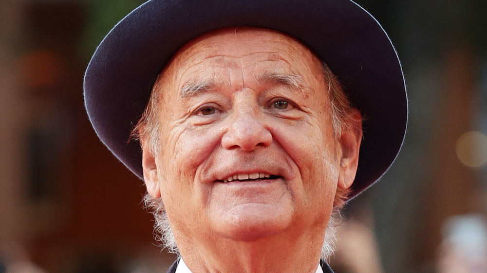 Bill Murray smiles wearing a dark blue brimmed hat