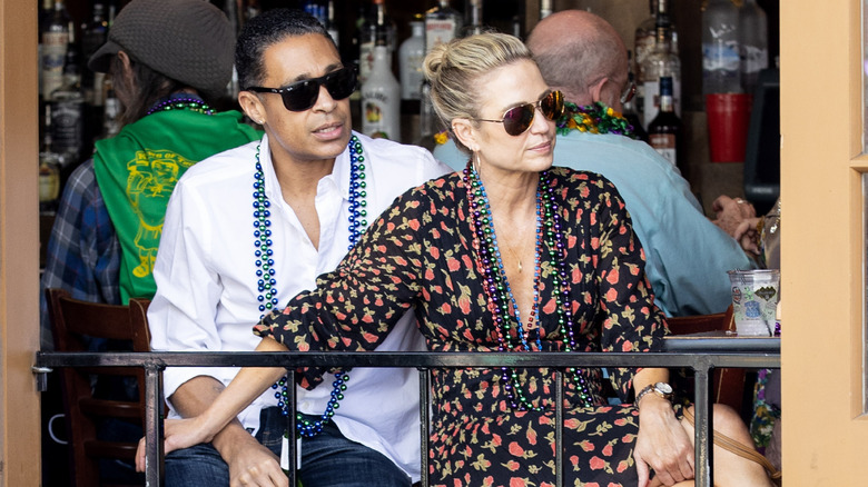 T.J. Holmes and Amy Robach seated on a balcony, wearing Mardi Gras beads and sunglasses.