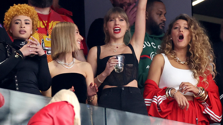 Blake Lively wearing red tracksuit and gold jewelry at the 2024 Super Bowl while standing beside Taylor Swift