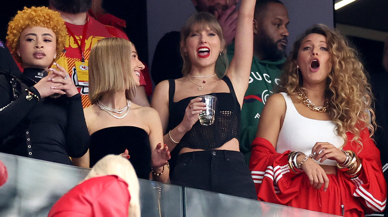 Blake Lively wearing red tracksuit and gold jewelry at the 2024 Super Bowl while standing beside Taylor Swift