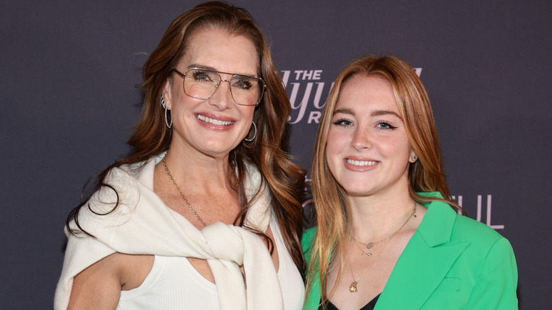 Brooke Shields wearing a white sweater tied around her shoulders and smiling next to daughter Rowan Henchy, who wears a green blazer.