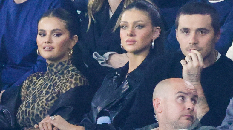 Selena Gomez and Brooklyn Beckham gaze sideways while Nicola Peltz stares ahead during a game.