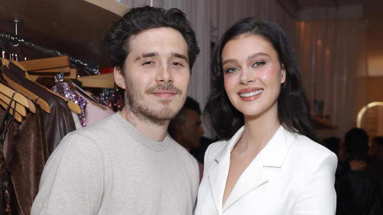Wearing beige and white outfits, Nicola Peltz and Brooklyn Beckham smile softly at the lens.