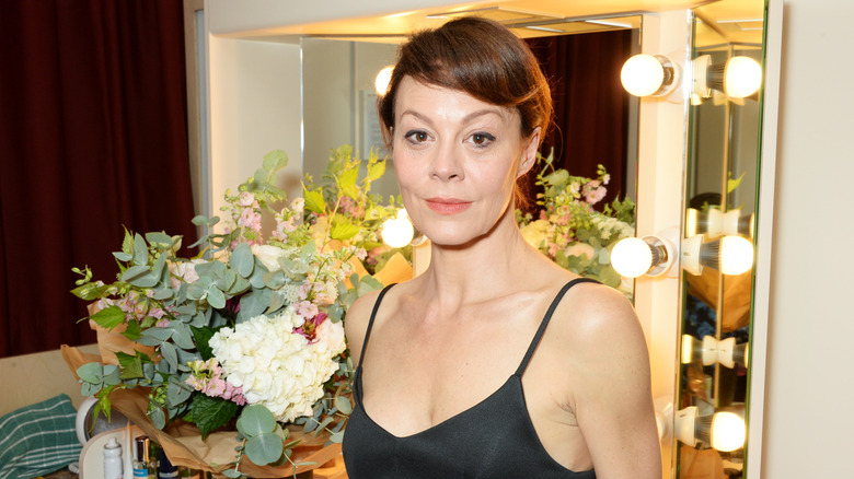 Helen McCrory wearing a black strappy nightgown while standing in front of a dressing mirror and bouquet of flowers