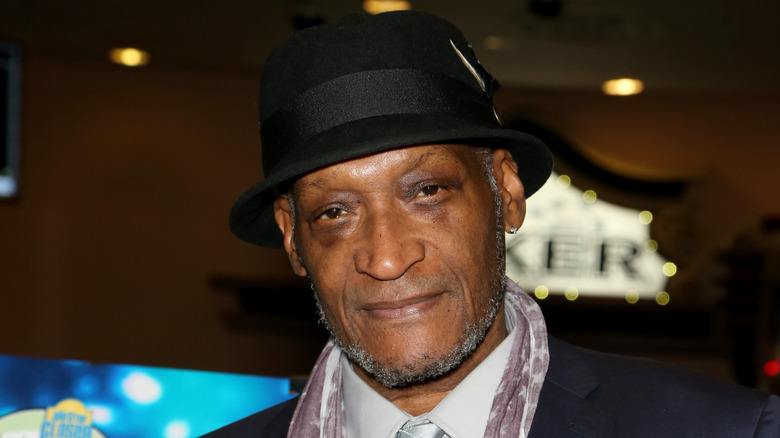 Tony Todd wearing a black hat, black jacket, white collared shirt and silver tie