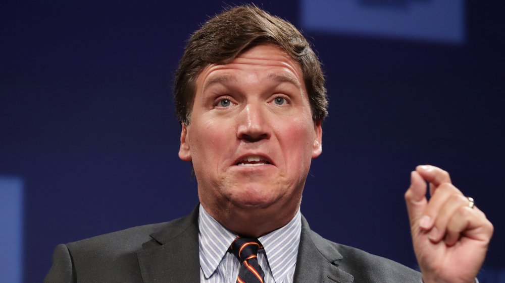 Tucker Carlson in a grey suit and striped shirt-and-tie combo, speaking with his hand raised