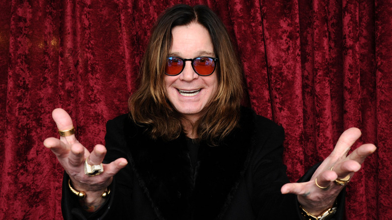 Ozzy Osbourne holding hands upward and outward with big grin