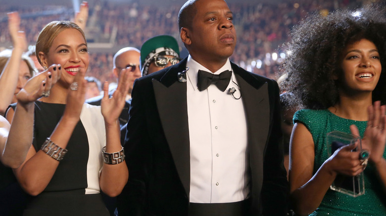 Jay-Z looking worried next to clapping Beyonce and Solange Knowles