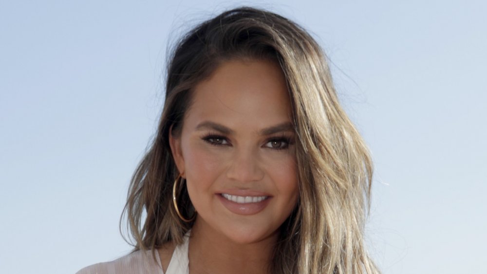 Chrissy Teigen in a white dress and gold hoop earrings, smiling on the beach