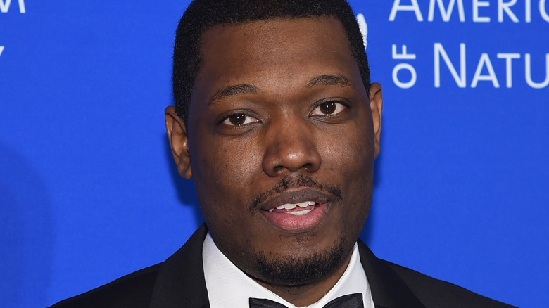 Michael Che in a black tux, posing at a museum event