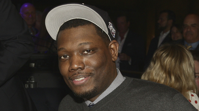 Michael Che in a gray sweater and black hat, posing with a small smile