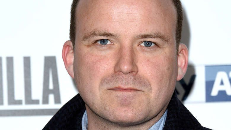Rory Kinnear in a black-and-blue checkered coat, posing with a neutral expression