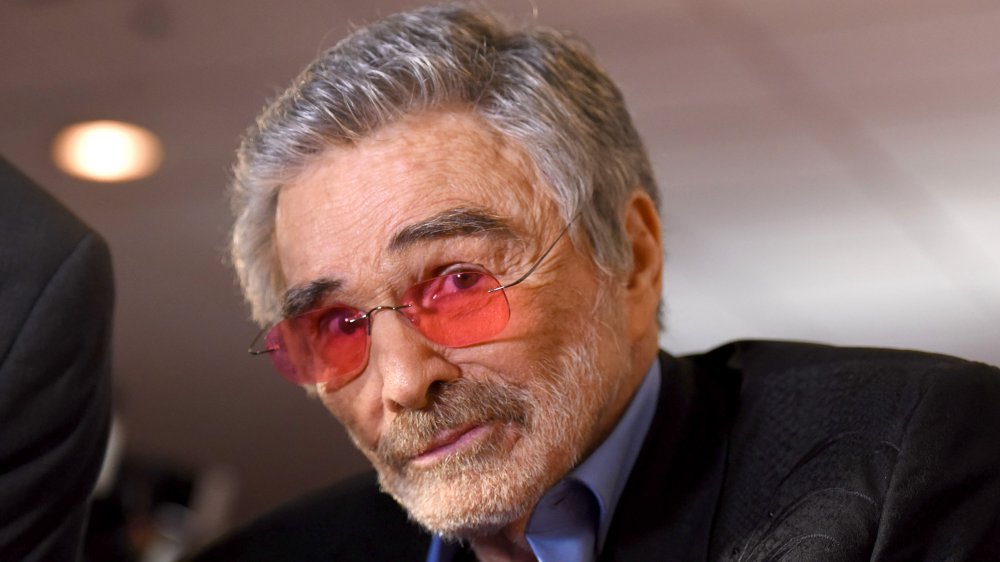 Burt Reynolds in a black blazer, blue shirt, and orange-tinted glasses, with a surprised expression